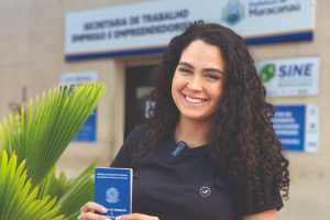 Foto de moça sorridente, segurando a carteira de trabalho, em frente ao Sine Municipal