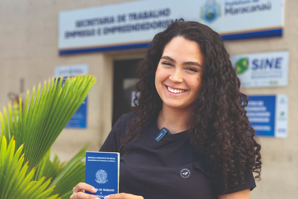 Foto de moça sorridente, segurando a carteira de trabalho, em frente ao Sine Municipal